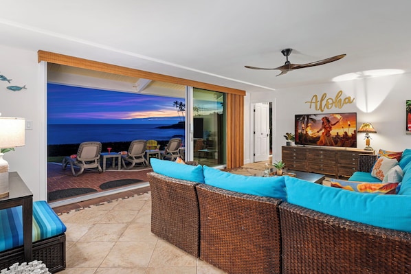 A living room with a view of the ocean, wicker furniture, and a ceiling fan.