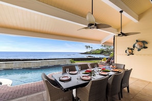 An outdoor dining area with a view of the ocean, a ceiling fan, and a table set for dining.