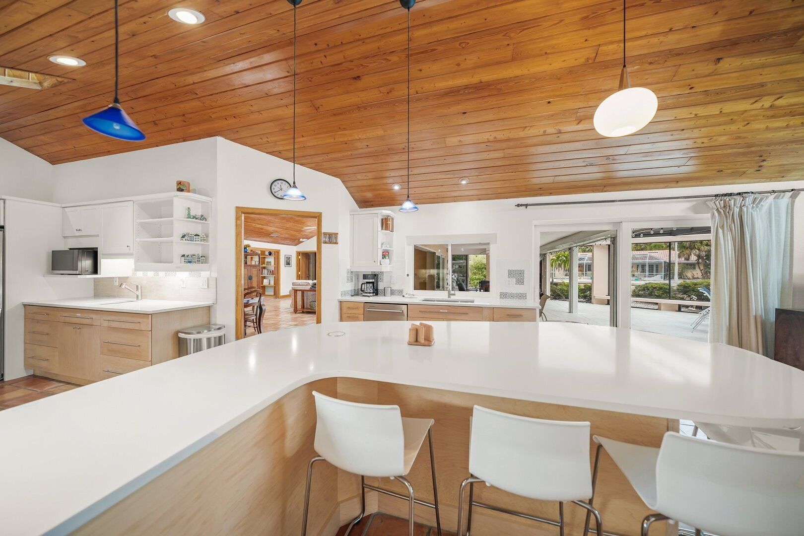 A modern kitchen with a large island, white bar stools, wooden ceiling, and a view of the outdoors.