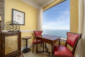 Una habitación con vista al océano, una mesa de madera, sillas rojas, un cuadro enmarcado en la pared y un objeto metálico decorativo sobre un pedestal.