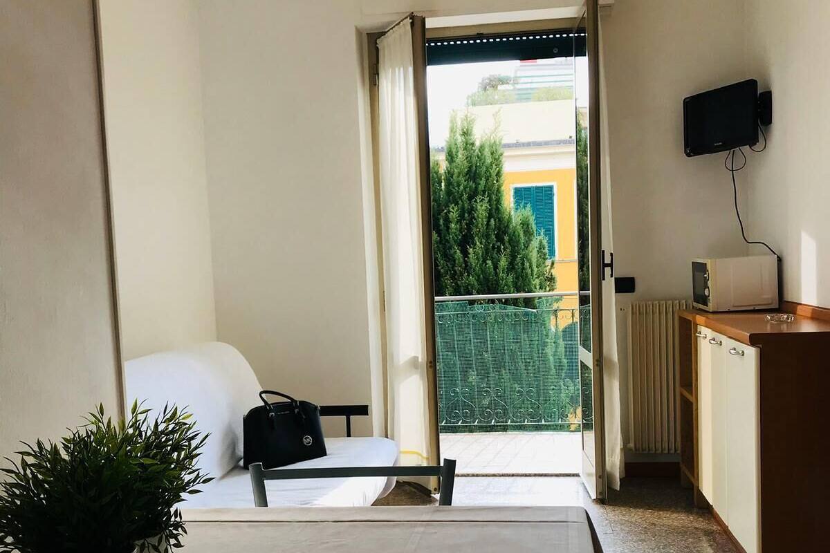 A hotel room with a bed, a small table, a potted plant, a desk with a computer, and a view of the outside balcony.