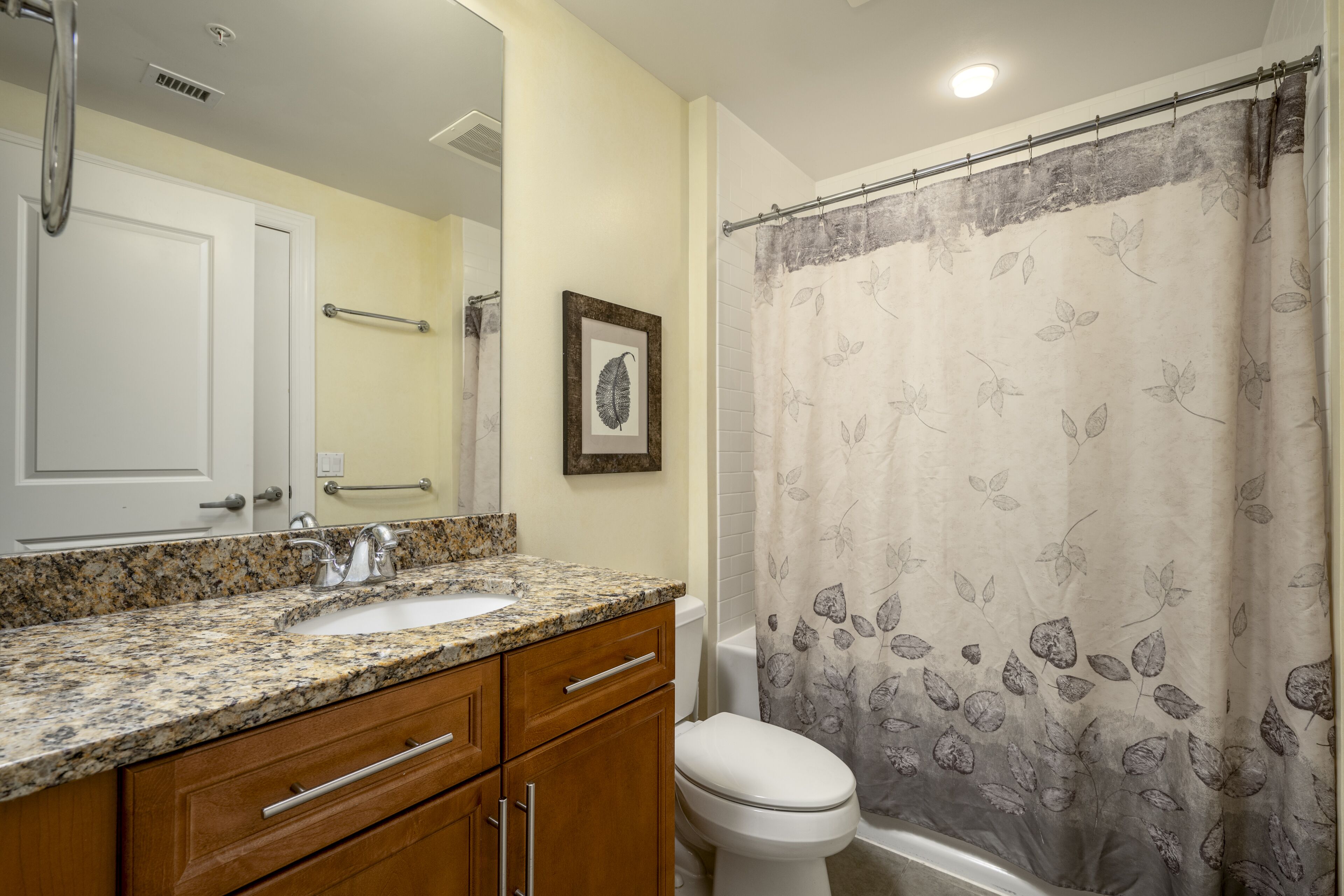 A bathroom with a granite countertop sink, wooden cabinets, a white toilet, and a shower with a leaf-patterned curtain.