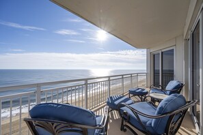 Un balcón con sillas azules acolchadas y una mesa, con vistas a una playa, el océano y un cielo despejado.