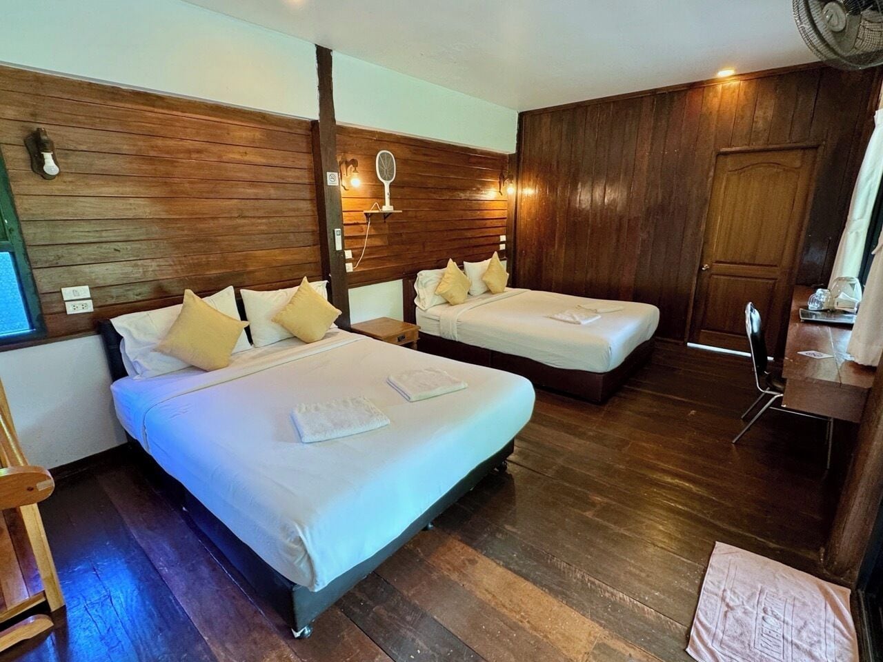 Habitación de hotel con dos camas, paneles de madera en las paredes, una mesita con una silla y un ventilador.