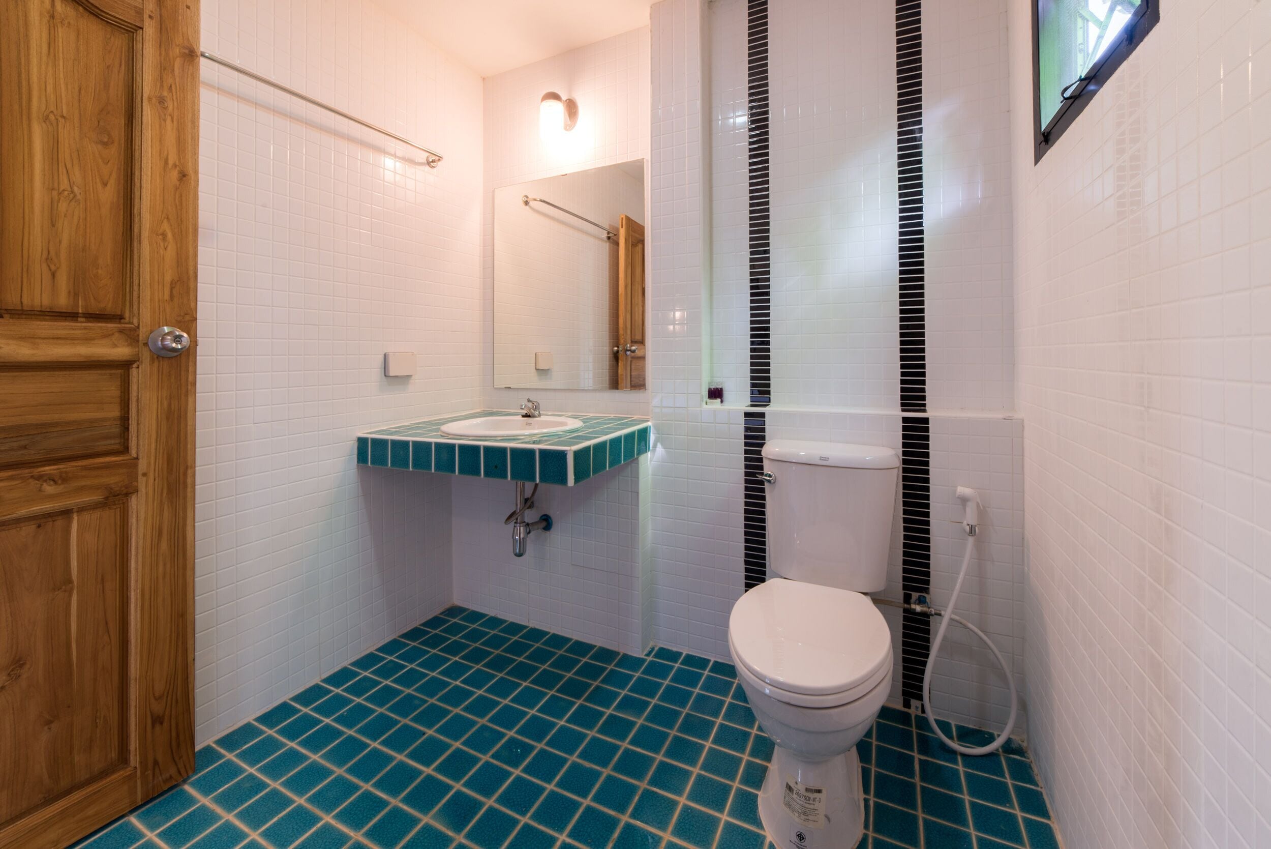 Une salle de bain avec une porte en bois, des murs recouverts de carreaux blancs, un sol carrelé de bleu, des toilettes blanches, un lavabo avec un plan de travail vert et un miroir au-dessus du lavabo.