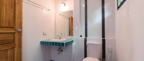 Une salle de bain avec une porte en bois, des murs recouverts de carreaux blancs, un sol carrelé de bleu, des toilettes blanches, un lavabo avec un plan de travail vert et un miroir au-dessus du lavabo.