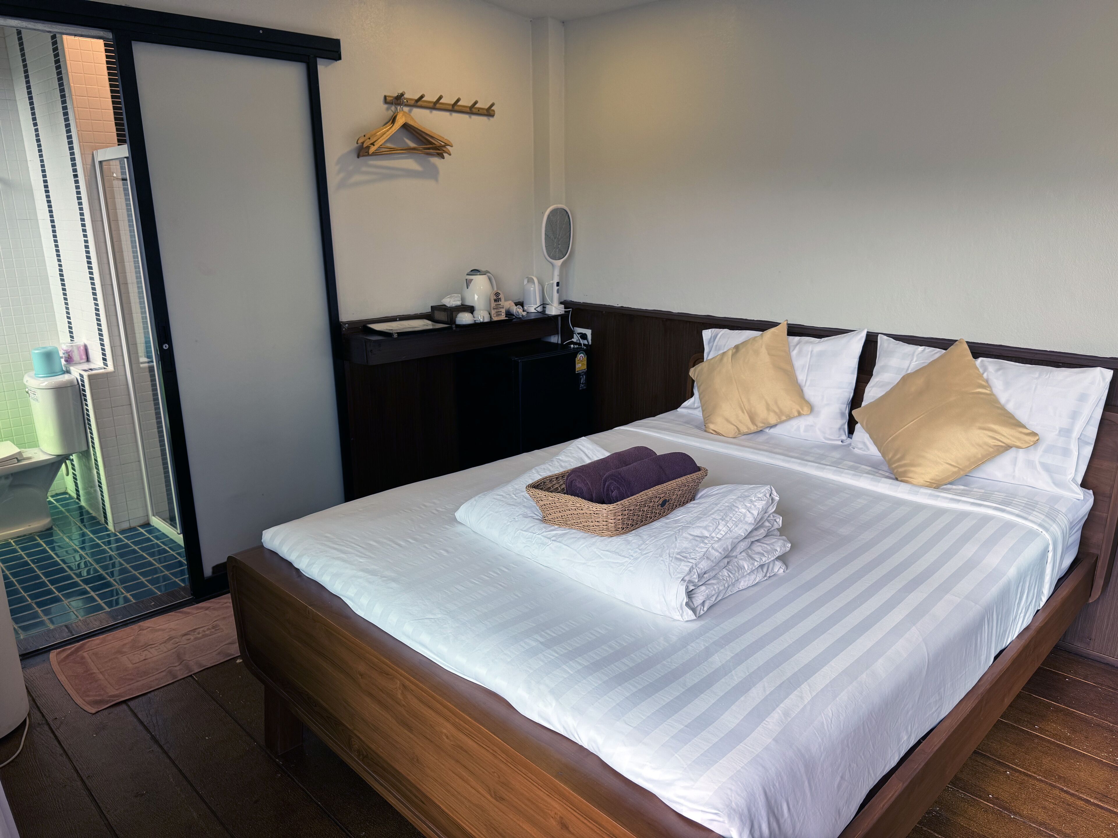 A neatly made bed with white linens, two pillows, a basket of folded towels, and a wooden headboard.