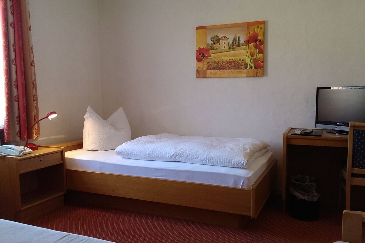 A hotel room with a single bed, a desk with a computer, a chair, a lamp, and a painting on the wall.