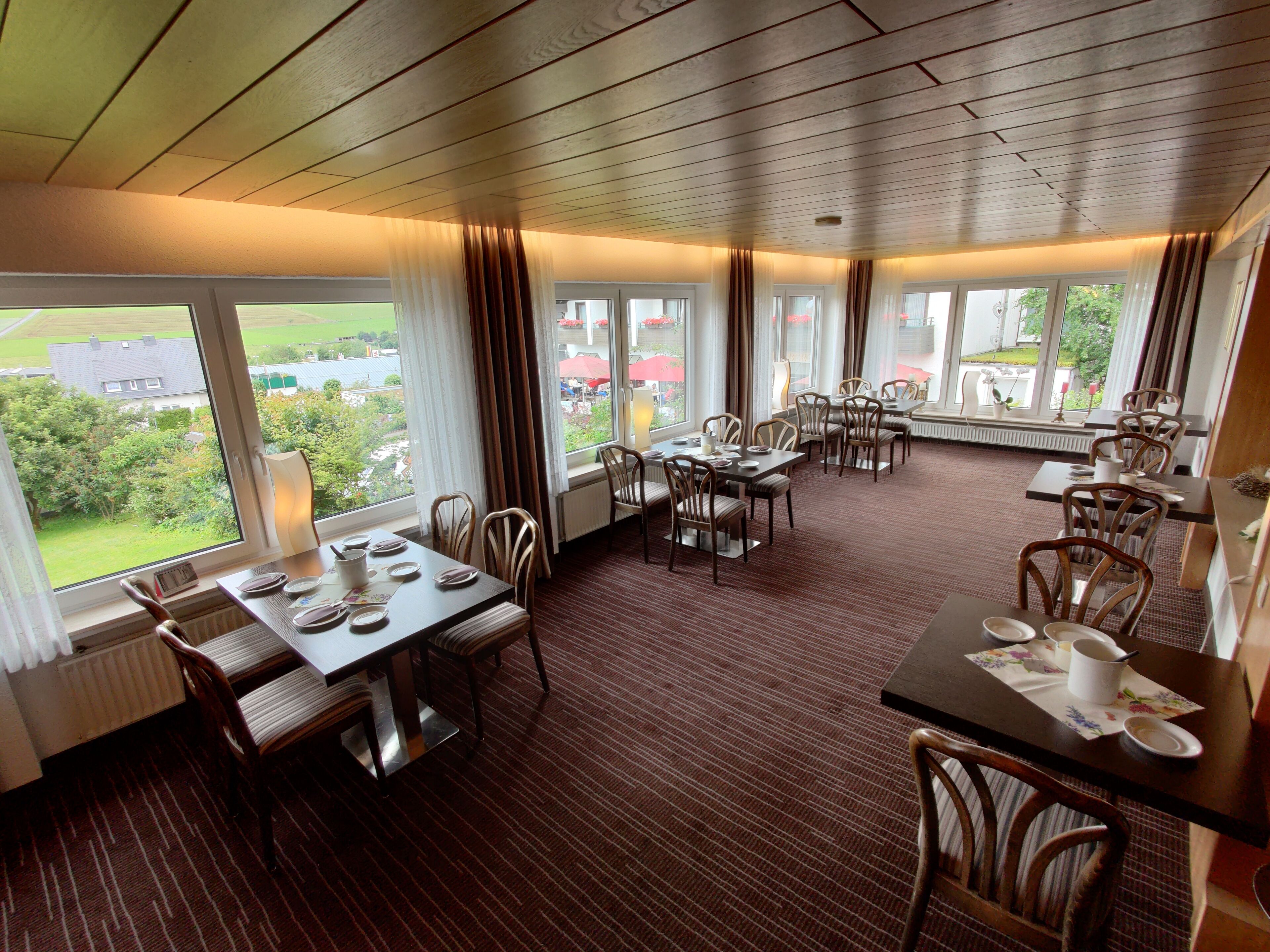Comedor de hotel con varias mesas y sillas, ventanas amplias con cortinas y vistas al exterior.