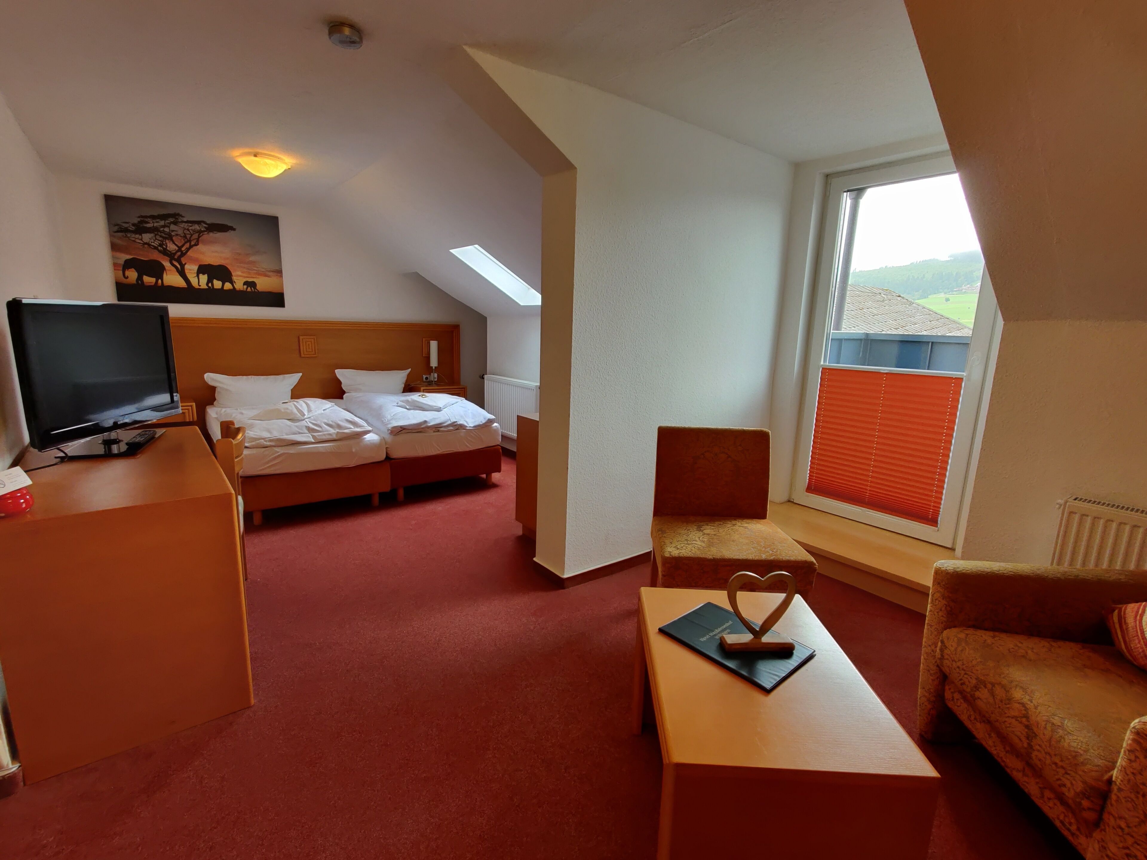 A hotel room with a bed, a desk with a TV, a chair, a small table with a heart-shaped decoration, and a painting of elephants on the wall.