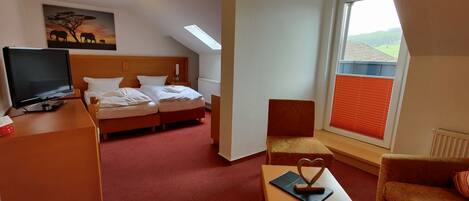 A hotel room with a bed, a desk with a TV, a chair, a small table with a heart-shaped decoration, and a painting of elephants on the wall.