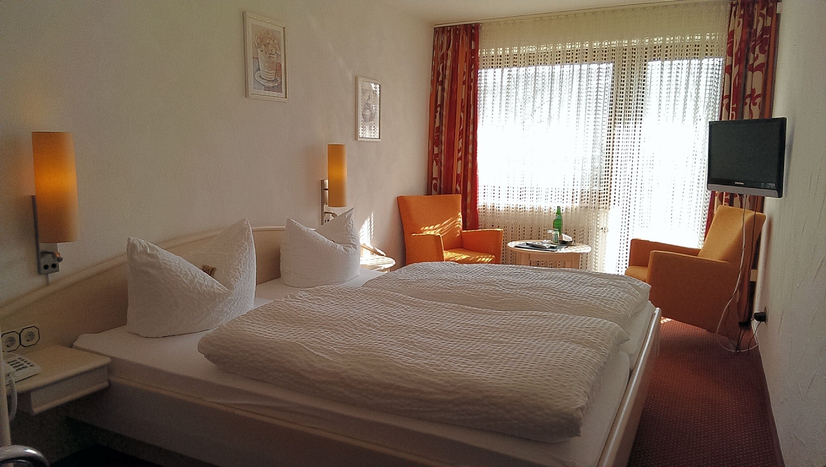 A hotel room with a large bed, two chairs, a small table with a bottle, and a TV on the wall.