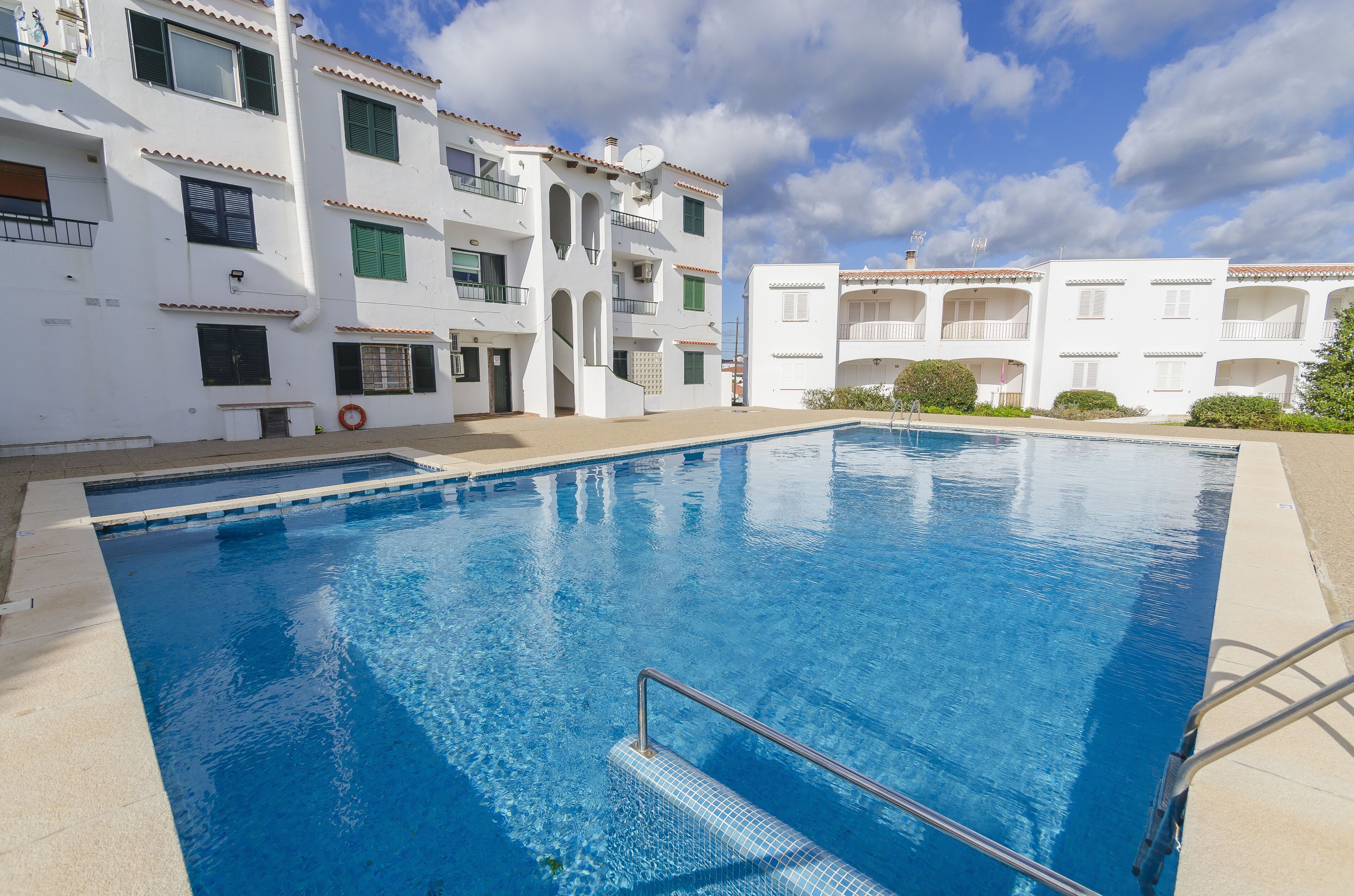 Una piscina con escalera metálica, rodeada de edificios blancos con balcones y contraventanas verdes.