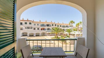 A balcony with wicker chairs and a table, overlooking a street with buildings and palm trees.