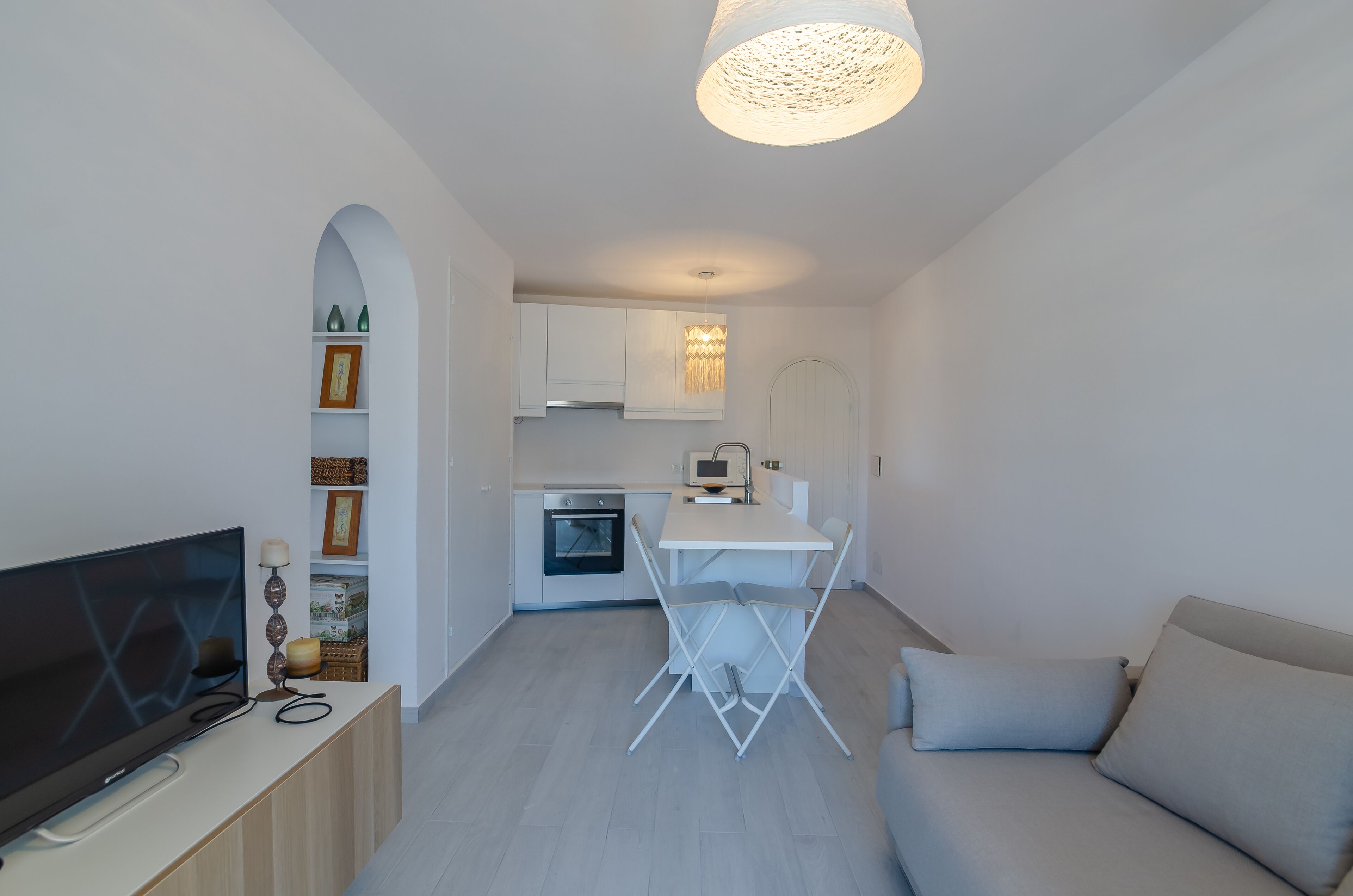A modern living room with a flat-screen TV, a sofa, and a dining table with chairs. There is a kitchen area with cabinets and a microwave.