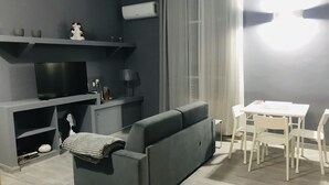 Moderni olohuone, jossa on harmaa sohva, ruokapöytä tuoleineen, taulutelevisio ja pieni hylly koriste-esineille.