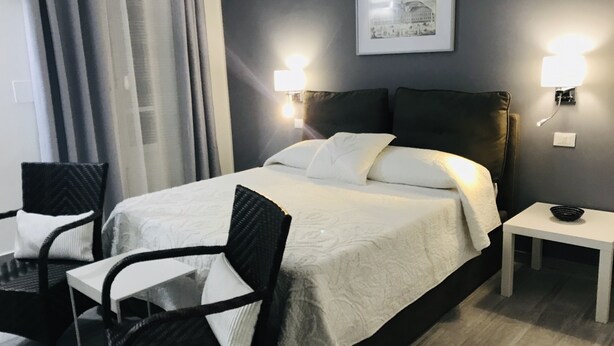 Hotellihuone, jossa on sänky, kaksi tuolia, yöpöytä ja seinään kiinnitetty valaisin.