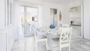 A white room with a round table, white chairs, a glass door leading outside, and a white shelving unit.