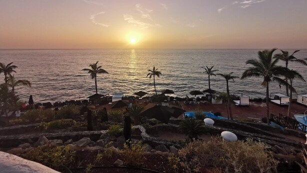 A beachside resort with palm trees, sun loungers, and a pool area during sunset.