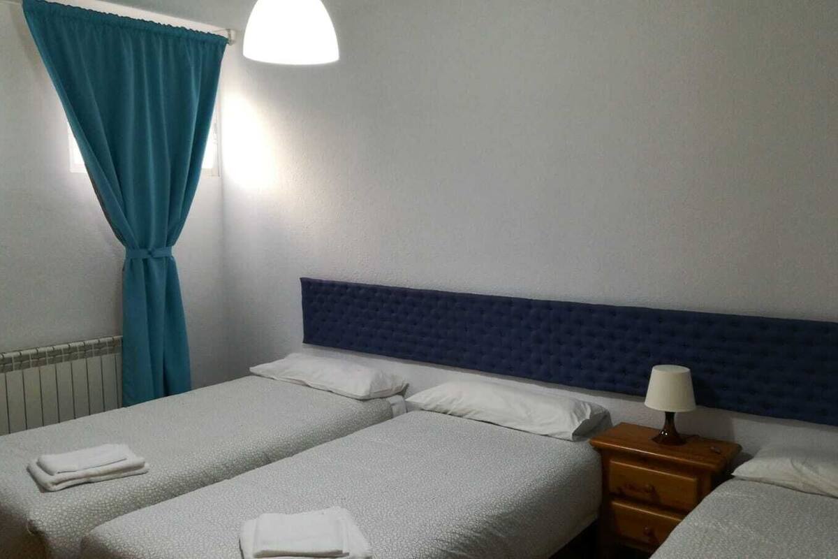 Habitación de hotel con dos camas, cabecera azul, mesita de noche con lámpara y ventana con cortinas.