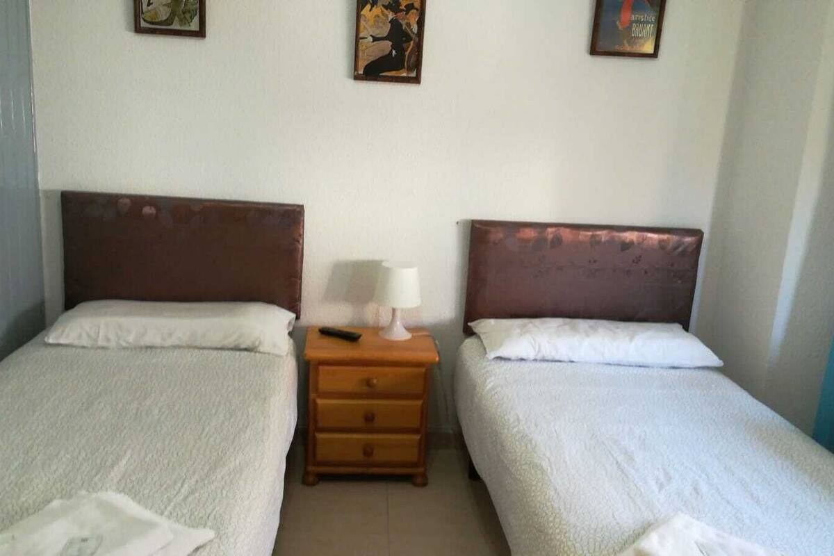 Una habitación con dos camas individuales, una mesita de noche de madera con lámpara y una pequeña mesa.