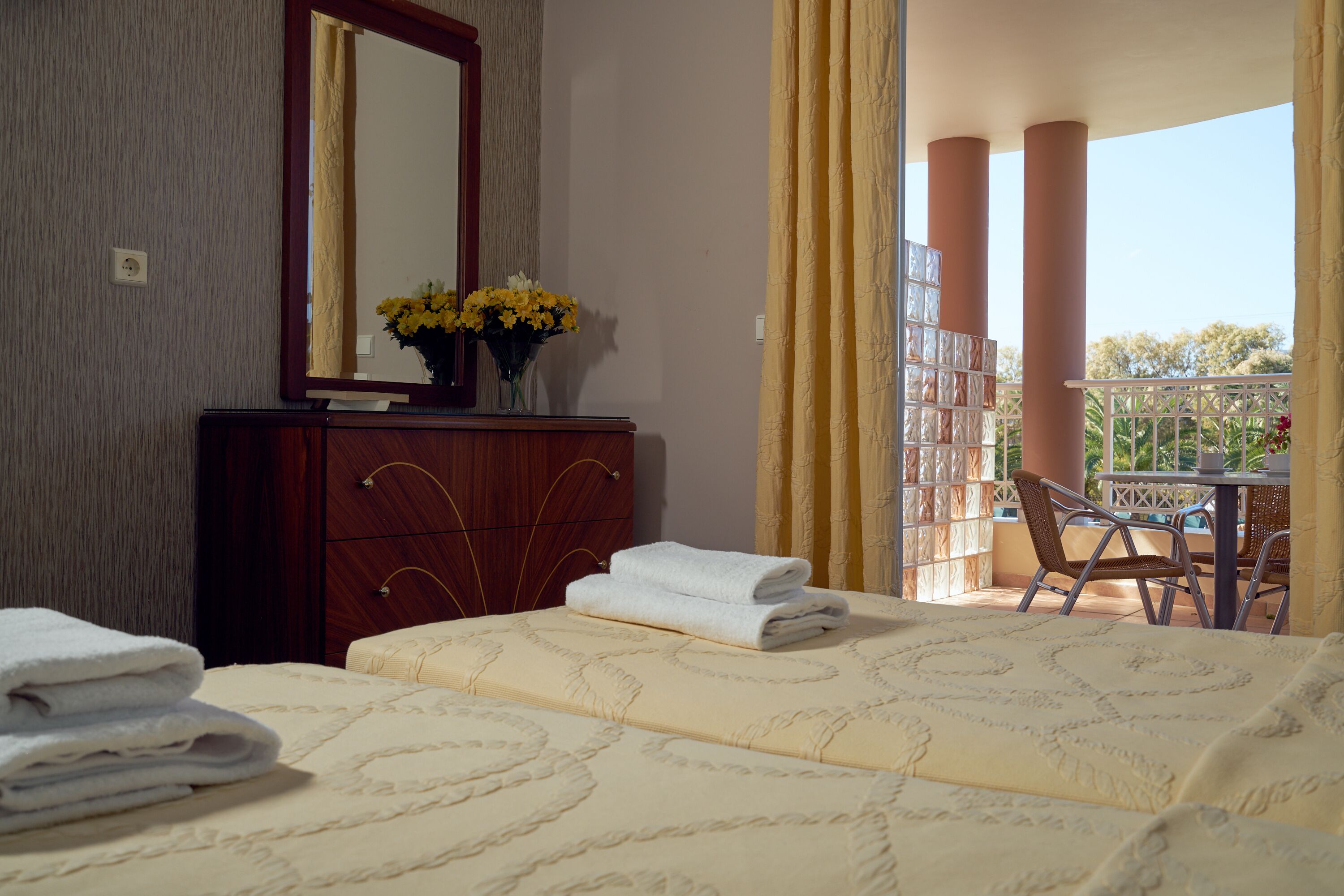 A hotel room with a bed, a dresser, a mirror, a vase with flowers, and a balcony with a table and chairs.