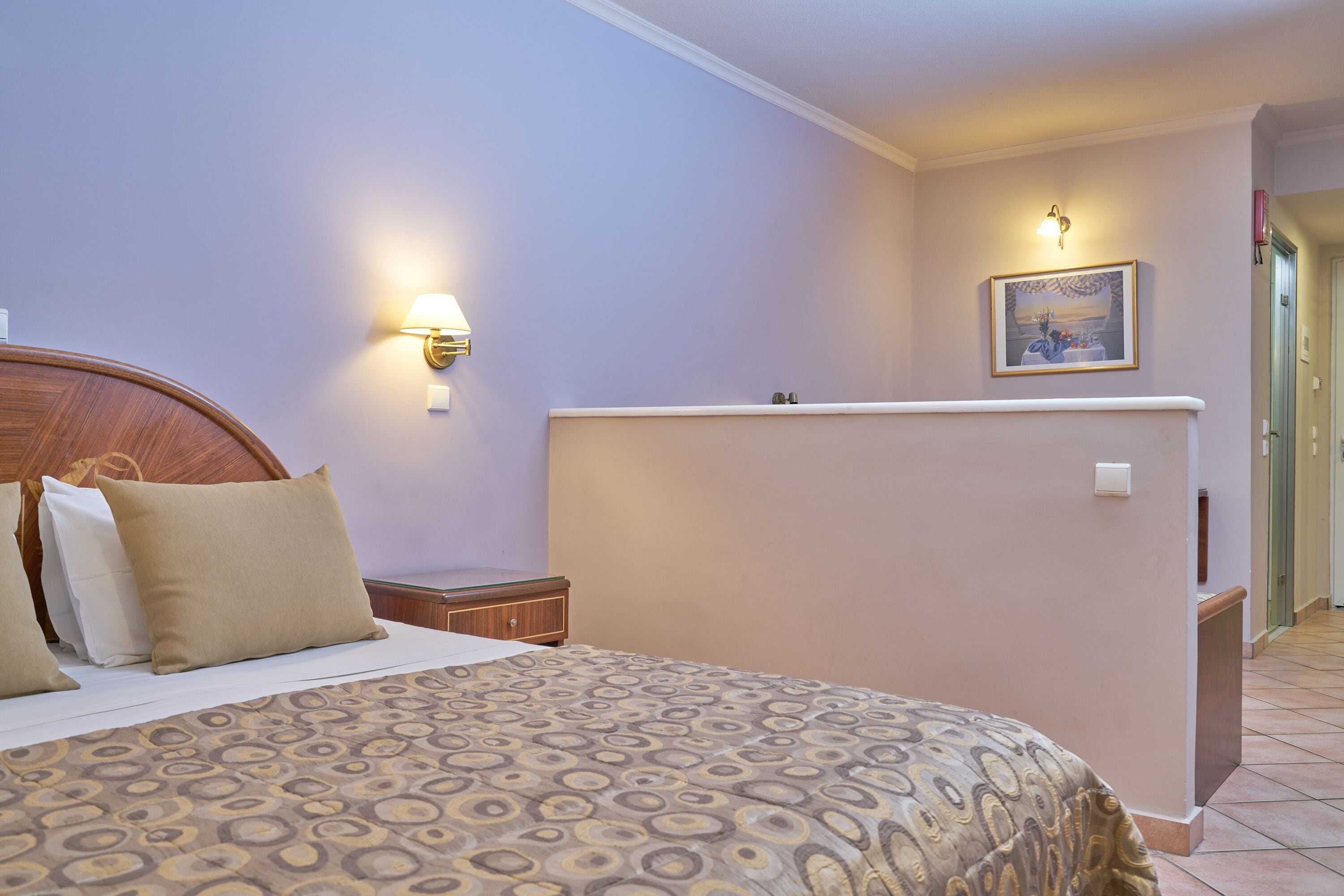 A hotel room with a bed, bedside table, wall-mounted lamp, and a painting.