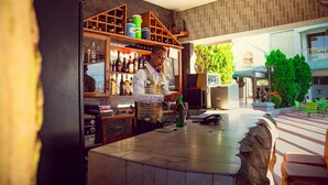 Una zona de bar con un bartender preparando una bebida, estantes con botellas y vista al área de asientos al aire libre.