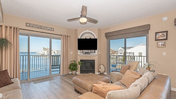 A spacious living room with a large sofa, a flat-screen TV, and a fireplace. There are large windows offering a view of the ocean and a balcony. - Upscale, stylish 3 bedroom condo with free WiFi, and outdoor pool, and a breathtaking view of the bay located in midtown and only two blocks from the beach! (Ocean City)
