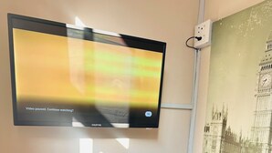A flat-screen TV mounted on a wall, a black chair, a desk, and a wall socket. - Karibu Guesthouse (Cape Town)