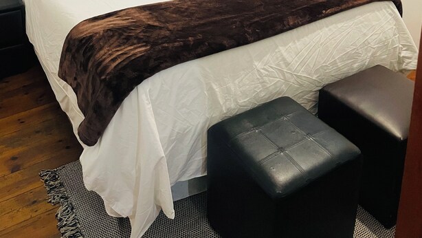 A neatly made bed with a grey headboard, a brown throw blanket, and a white pillow. There is a small black ottoman beside the bed. - Karibu Guesthouse (Cape Town)