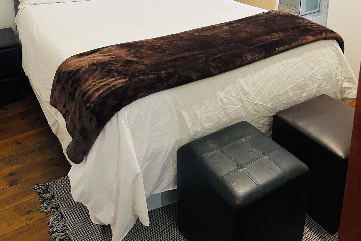 A neatly made bed with a grey headboard, a brown throw blanket, and a white pillow. There is a small black ottoman beside the bed.