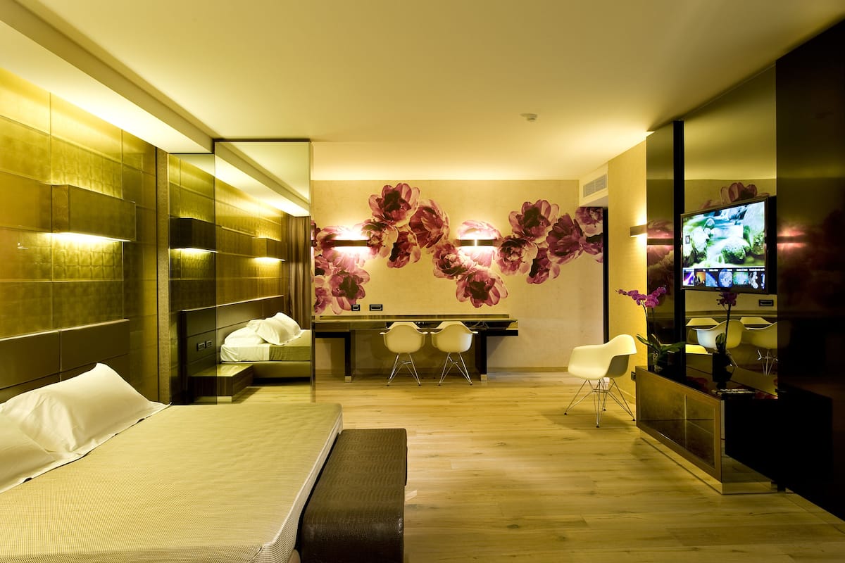 A modern hotel room with a large bed, a desk with two chairs, a television, and a decorative wall with floral patterns.