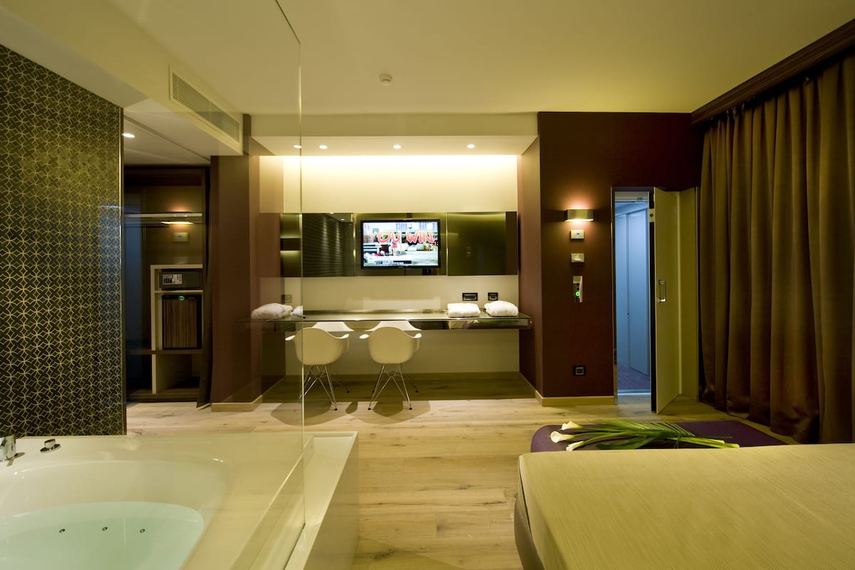 A modern hotel room with a large bathtub, a small kitchen area with a TV, and a separate dining space with two chairs.