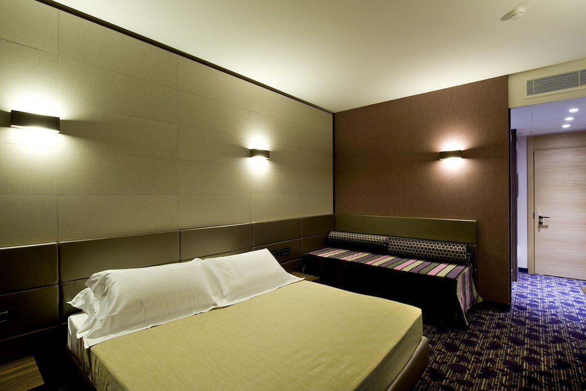 A hotel room with a large bed, a smaller sofa bed, and a wall-mounted light fixture.