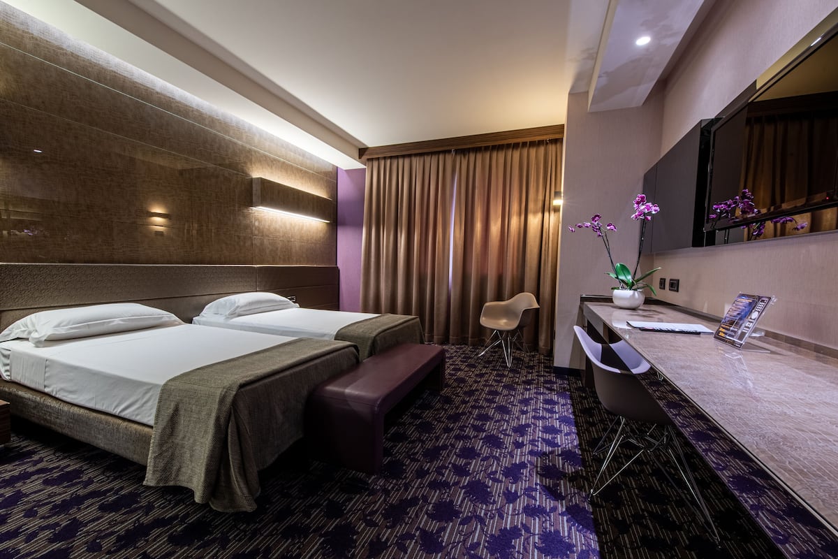 A hotel room with a large bed, a desk, a chair, a bench, and a potted orchid.