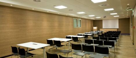 A conference room with rows of tables and chairs, a projector screen, and a wooden wall.