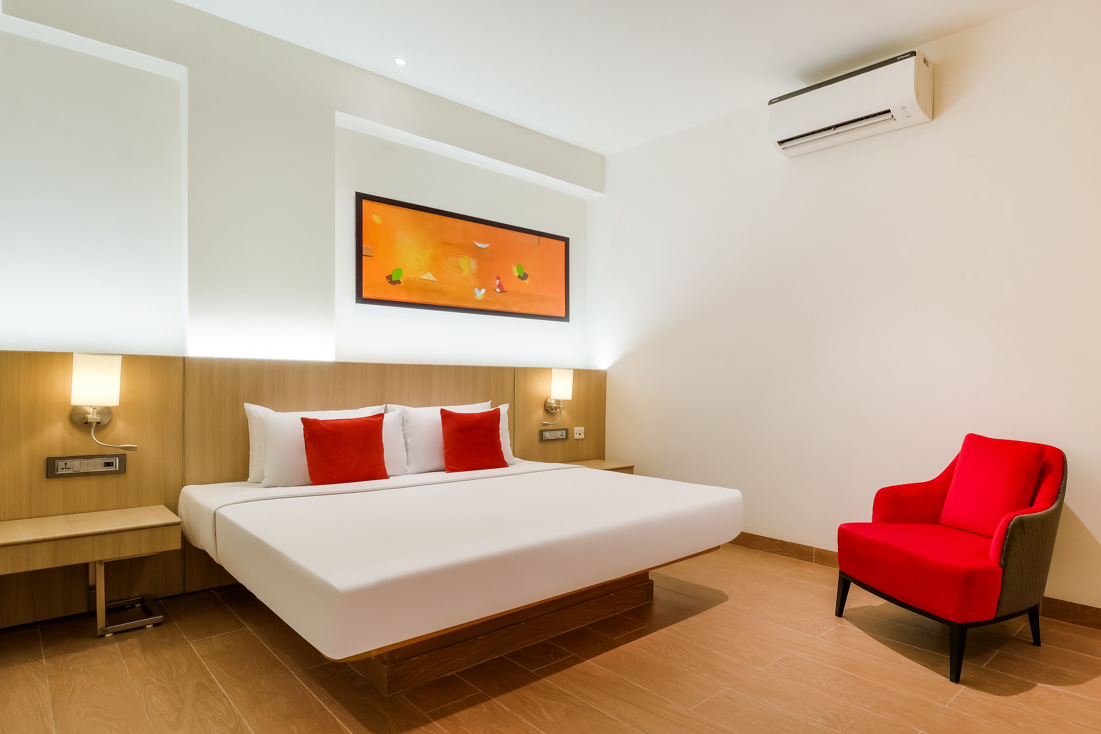 Une chambre d’hôtel avec un grand lit, un fauteuil rouge, un climatiseur fixé au mur et un tableau au-dessus du lit.