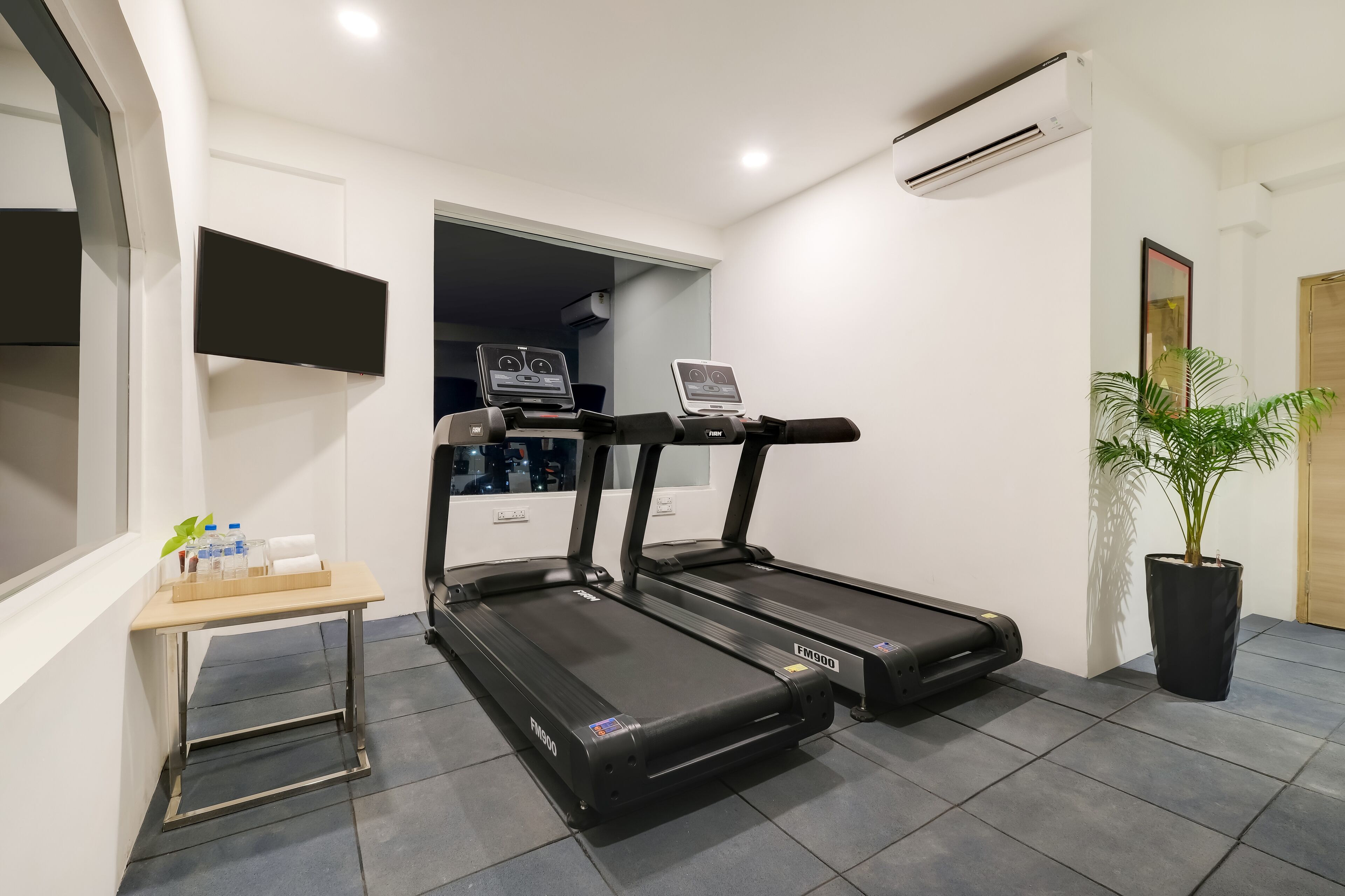 Un gimnasio en casa con dos cintas de correr, una mesita con botellas de agua y una planta en maceta.