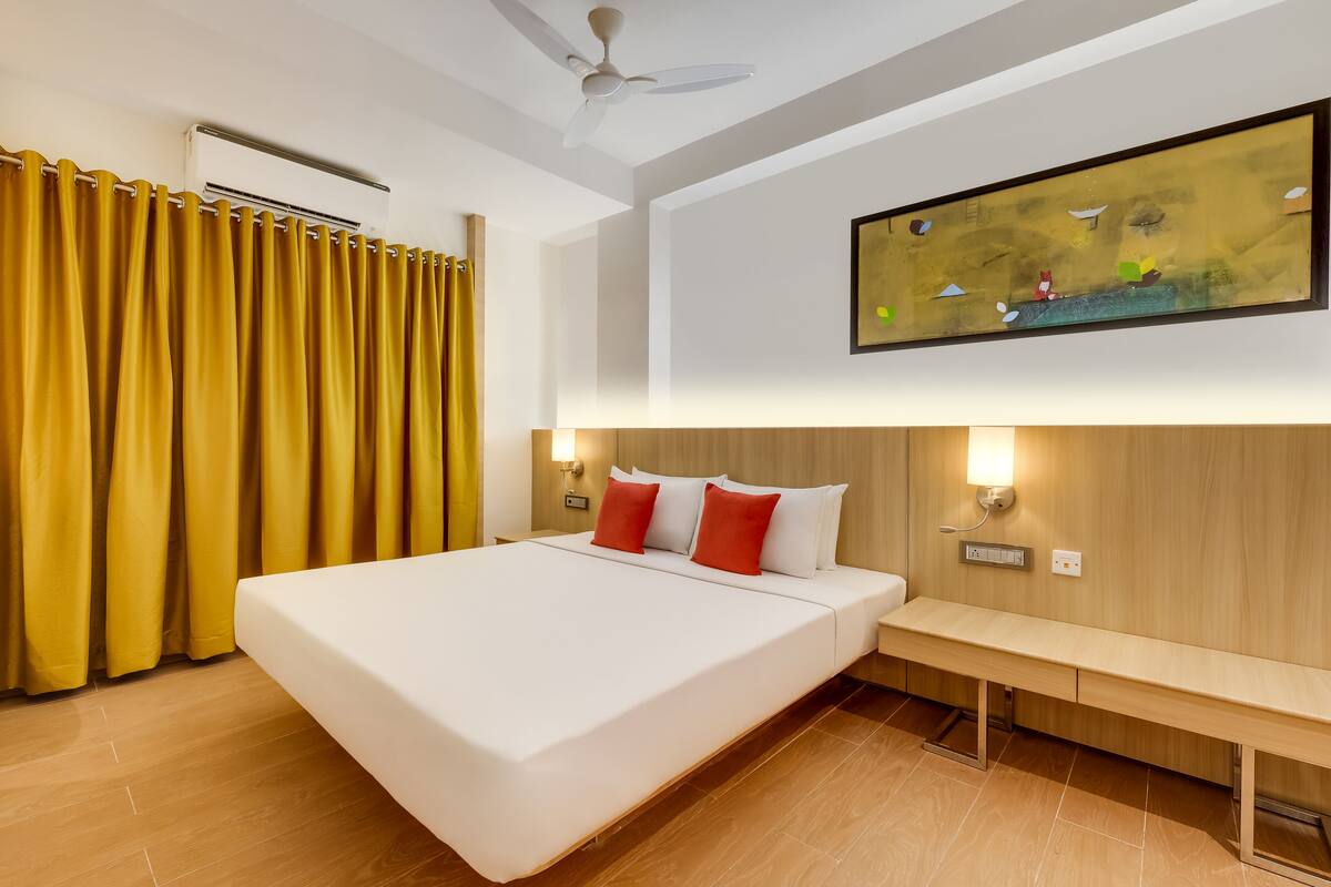 Une chambre d’hôtel avec un grand lit, des rideaux jaunes, un ventilateur de plafond, une lampe fixée au mur et un tableau.