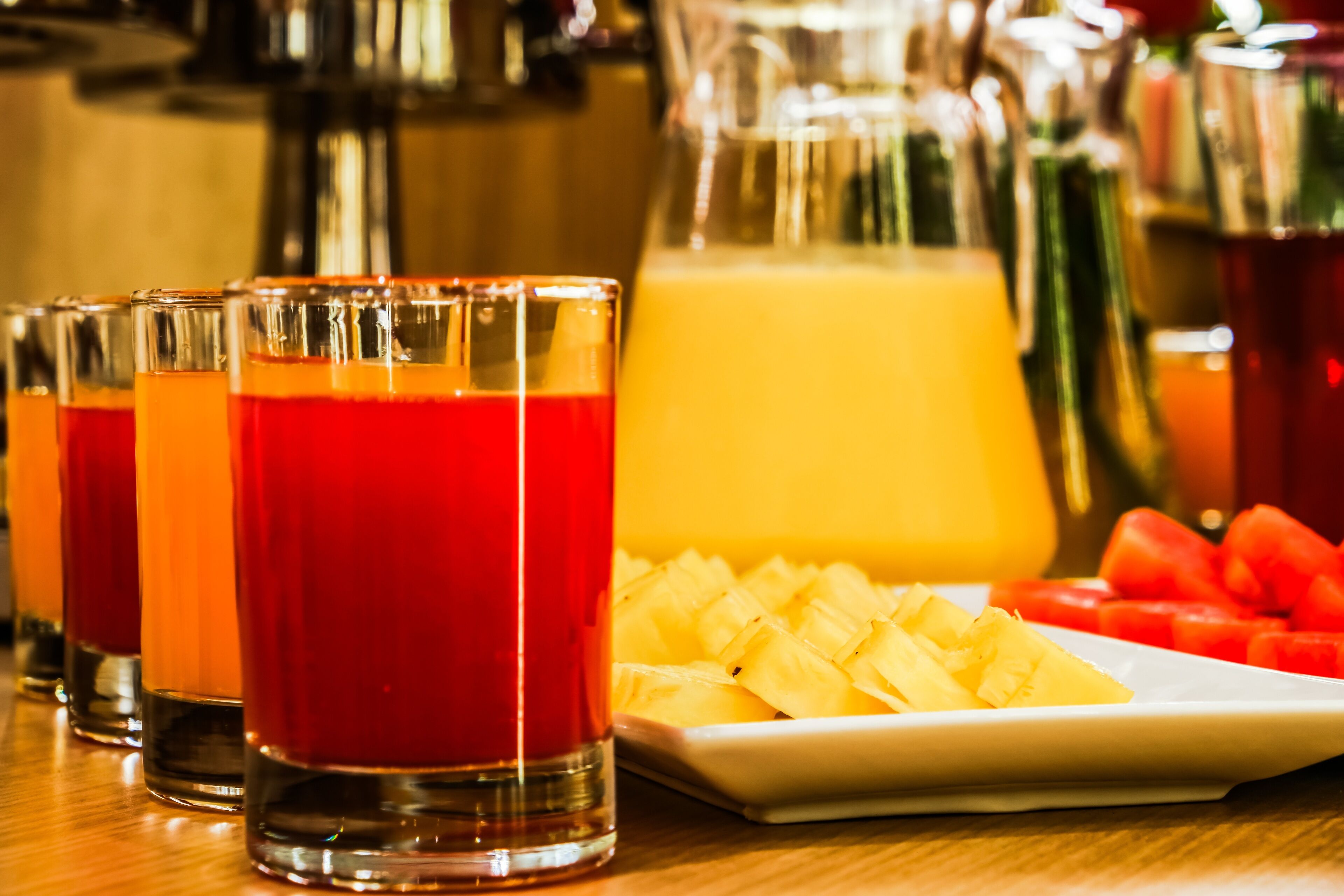 A bar setting with multiple glasses of red beverage, a pitcher of orange juice, and a plate of pineapple chunks.