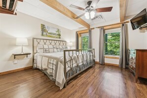 A bedroom with a bed, a ceiling fan, a painting, a window with curtains, and a TV.