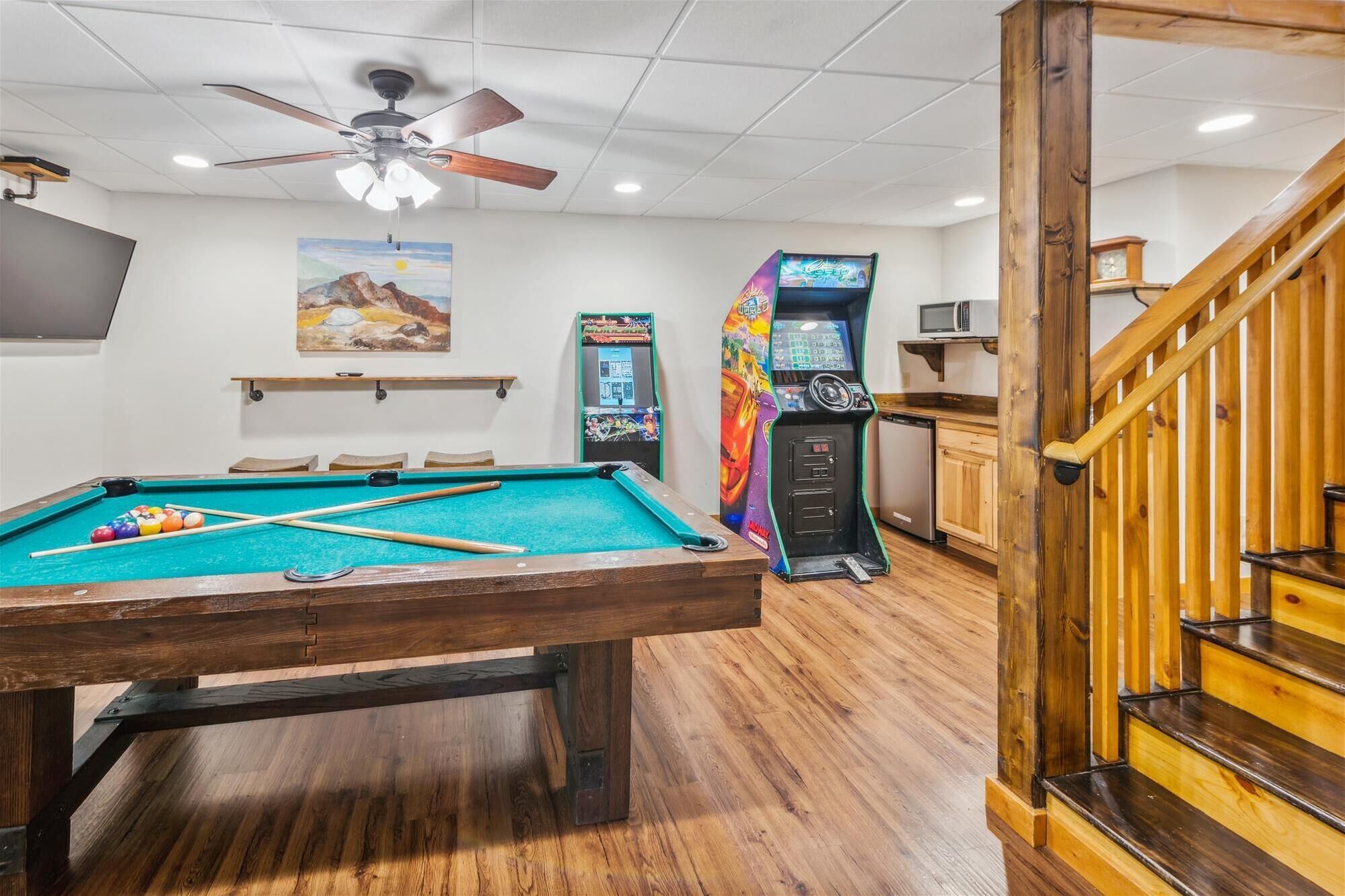 A pool table with pool cues, a ceiling fan, arcade games, and a wooden staircase.
