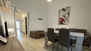 Un soggiorno moderno con zona pranzo, caratterizzato da un divano, un tavolino da caffè e un tavolo da pranzo con sedie. Si nota un quadro appeso alla parete e una porta che conduce a un'altra stanza.