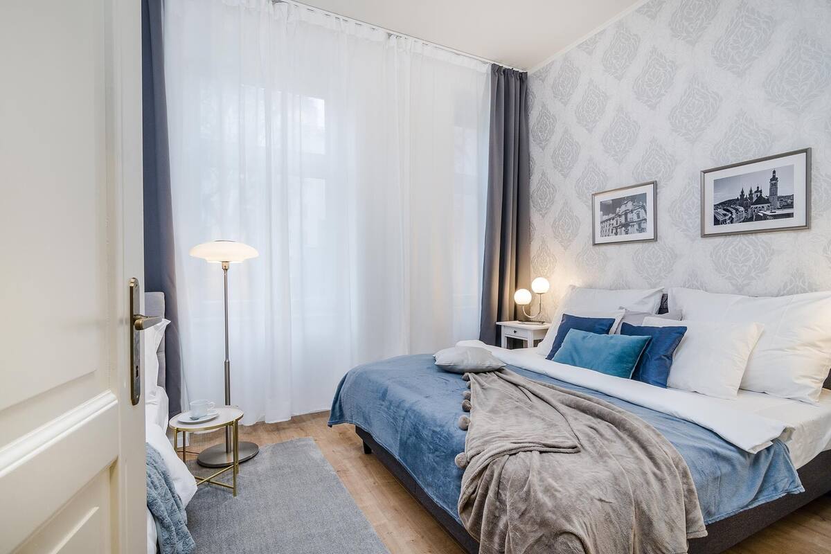 Ein Schlafzimmer mit einem Bett, einem Nachttisch mit Lampe, einem kleinen runden Tisch mit Tasse und Untertasse sowie einem Fenster mit durchsichtigen Vorhängen.