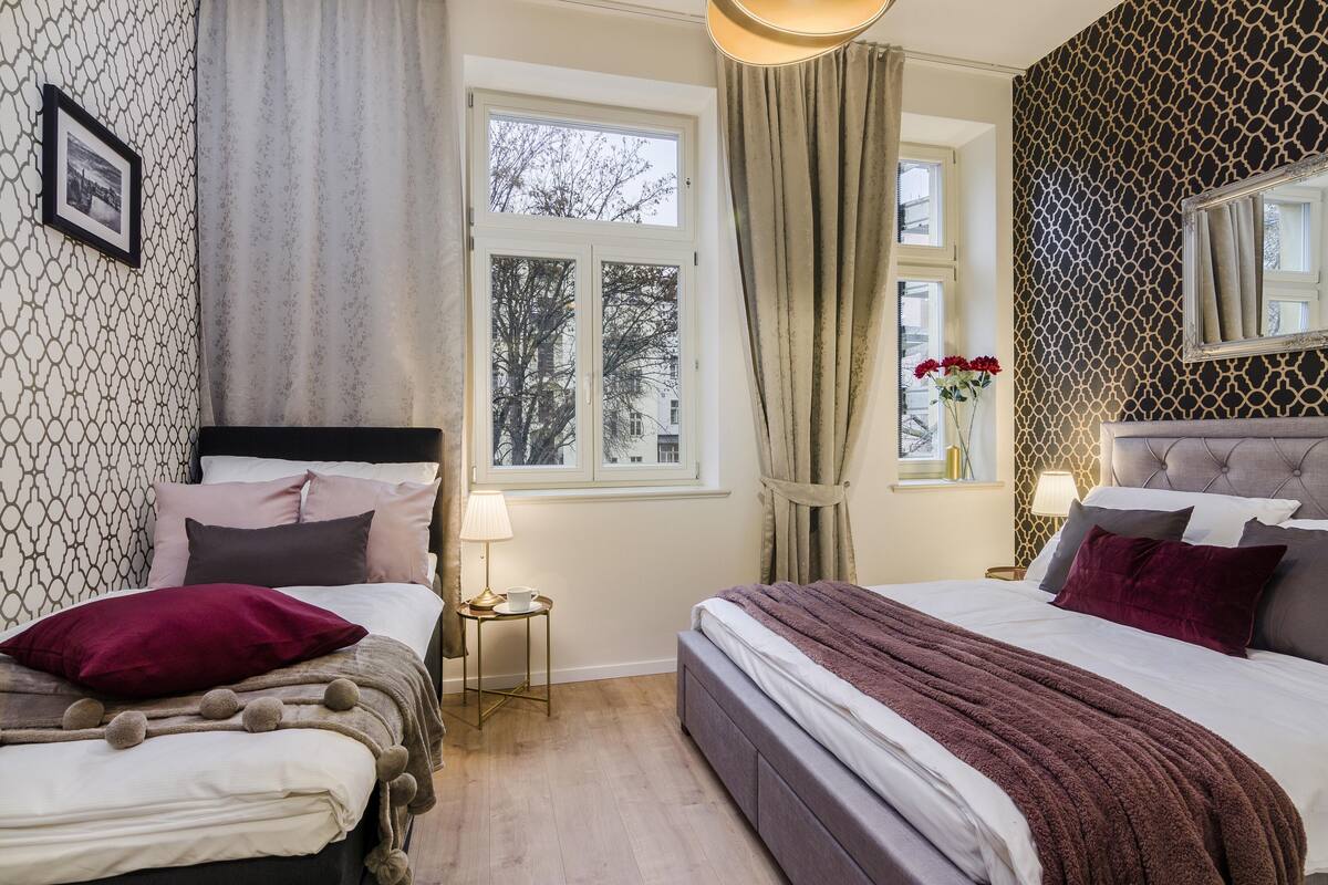 Ein Schlafzimmer mit zwei Betten, einem Fenster mit Vorhängen, einem Nachttisch mit Lampe und einer Wand mit geometrischem Muster.