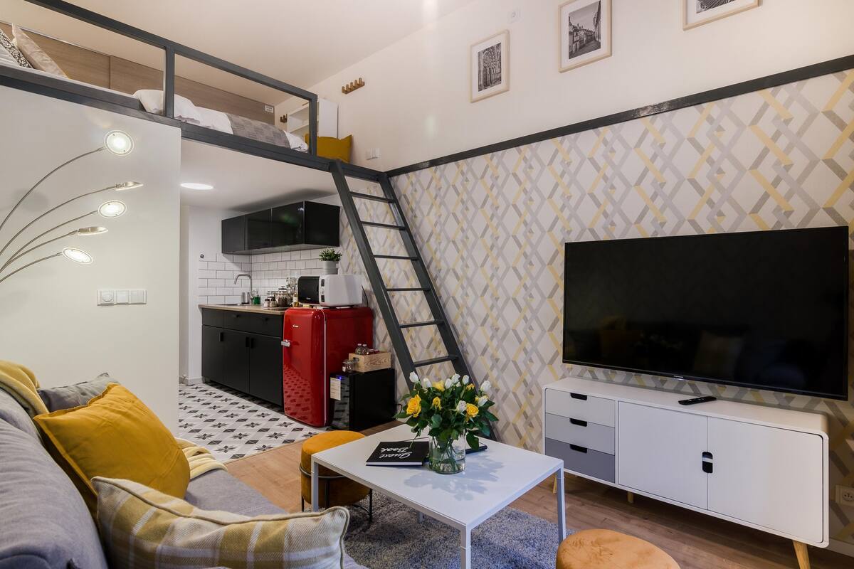 Ein kompakter Wohnraum mit einer Couch, einem Couchtisch, einem Fernseher, einer Küchenzeile und einer Wendeltreppe zu einem Erkerzimmer.