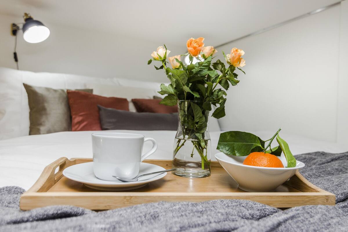 Ein ordentlich gedecktes Frühstücksbrett mit einer Tasse, einem Blumenstrauß und einer Obstschale auf einem Bett.