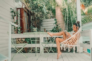 A person relaxing in a hammock chair on a porch with a table and chairs, surrounded by lush greenery and a wooden fence.