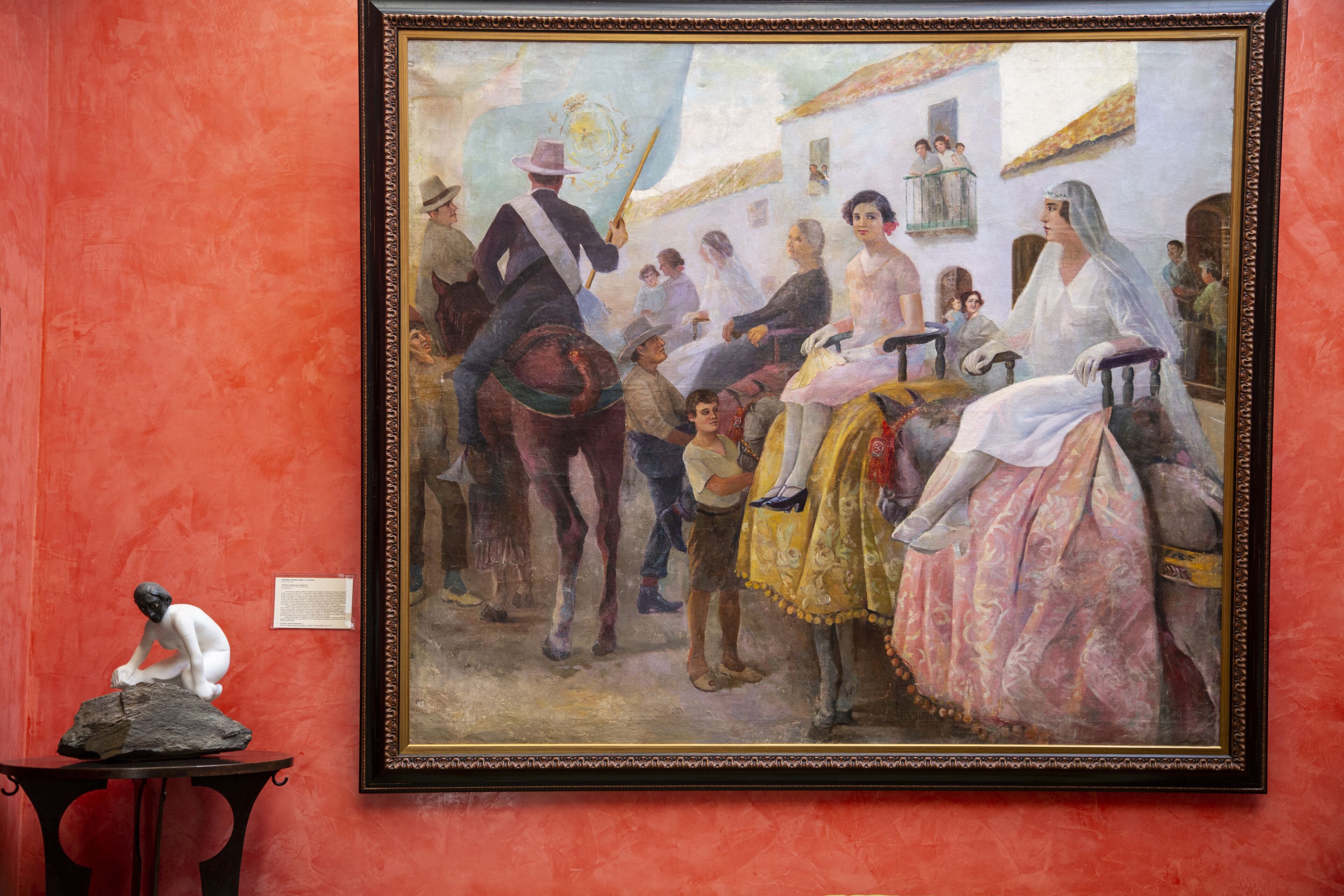 A framed painting of a historical scene with figures in period attire, a table with a sculpture, and a red wall.